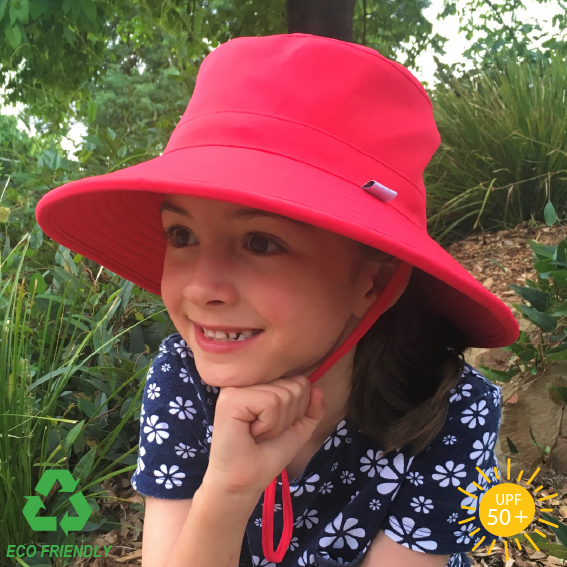 A child wearing a watermelon coloured Eco Friendly bucket hat with a wide brim, sitting outdoors with a garden and sunlight in the background.